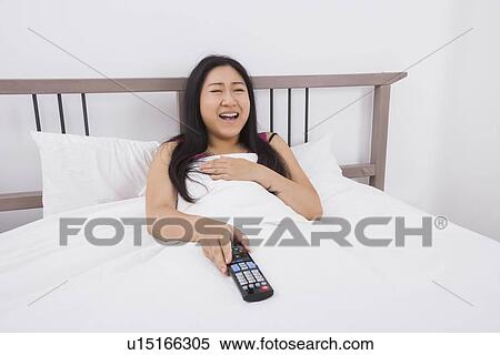 Stock Photography - Happy woman watching TV in bed. Fotosearch