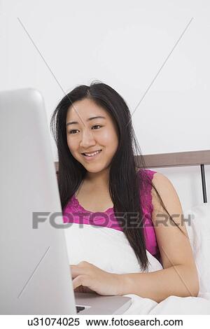 Stock Photography - Happy young woman using laptop on bed. Fotosearch