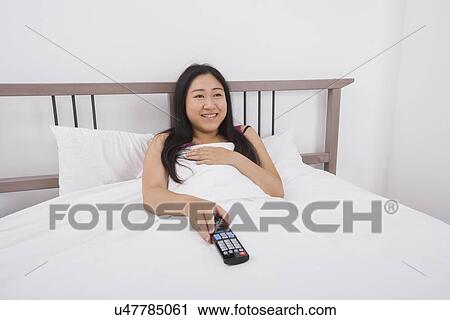 Stock Image - Happy young woman watching TV in bed. Fotosearch