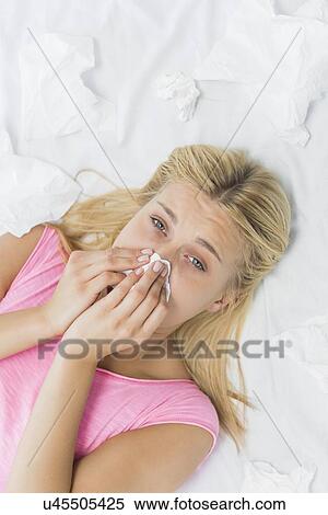 Stock Photography - High angle portrait of young woman blowing nose in tissue paper while lying on bed. Fotosearch