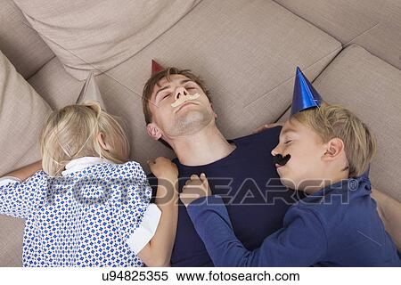 Stock Photography - High angle view of children and father with artificial mustache and party hat sleeping on sofa bed. Fotosearch