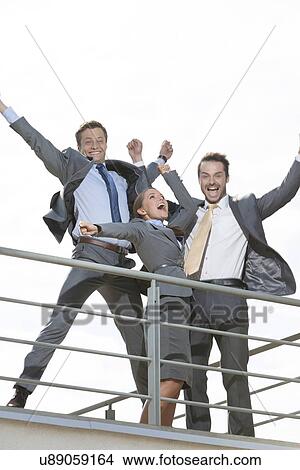 Picture - Low angle view of excited businesspeople with arms raised standing on terrace against clear sky. Fotosearch
