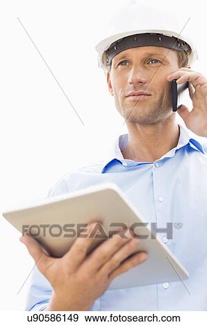 Stock Photo - Low angle view of male architect with digital tablet using cell phone against sky. Fotosearch