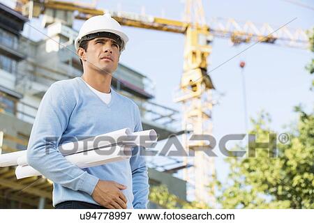 Stock Photo - Male architect holding rolled up blueprints while standing at construction site. Fotosearch