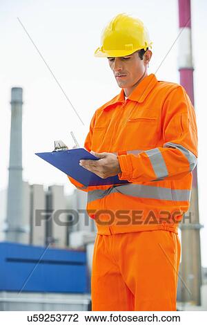Stock Image - Male architect in protective workwear writing on clipboard at site. Fotosearch