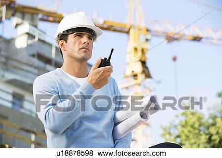 Stock Photo - Male architect with blueprints using walkie-talkie at construction site. Fotosearch