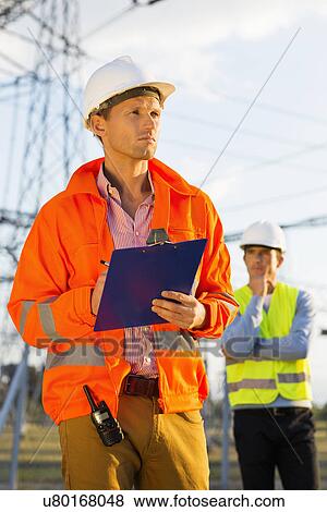 Male architect with clipboard working at site while coworker standing in background View Large Photo Image Stock Photo - Male architect with clipboard working at site while coworker standing in background. Fotosearch