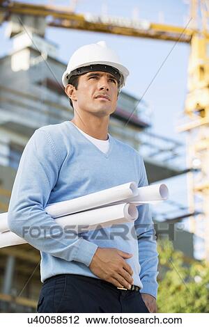 Stock Image - Male architect with rolled up blueprints standing at construction site. Fotosearch