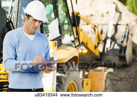 Picture - Male architect writing on clipboard against earthmover at construction site. Fotosearch