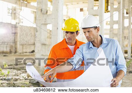 Stock Image - Male architects discussing over blueprint at construction site. Fotosearch