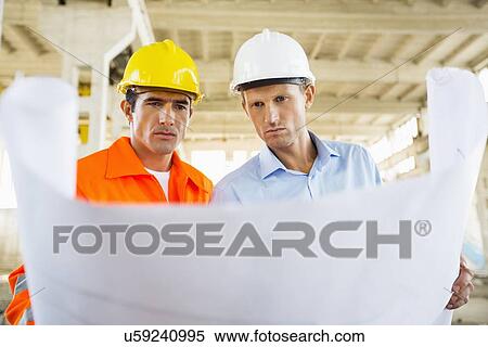 Stock Photography - Male architects reviewing blueprint at construction site. Fotosearch