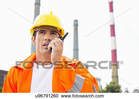 Stock Image - Male construction worker wearing reflective workwear communicating on walkie-talkie at site. Fotosearch
