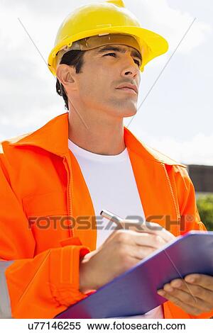 Stock Photography - Male construction worker with clipboard looking away outside. Fotosearch