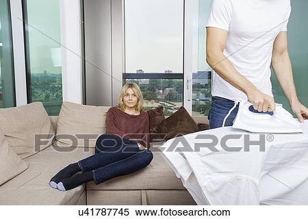 Stock Photography - Man ironing shirt while woman sitting on sofa at home. Fotosearch