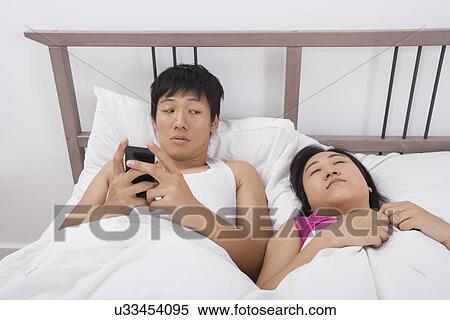 Stock Photography - Man using cell phone while looking at woman sleeping in bed. Fotosearch