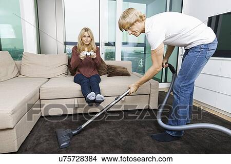 Picture - Man vacuuming while woman play video game in living room at home. Fotosearch