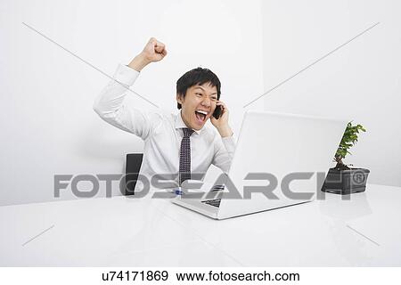 Stock Photo - Mid adult businessman cheering on cell phone at desk in office. Fotosearch