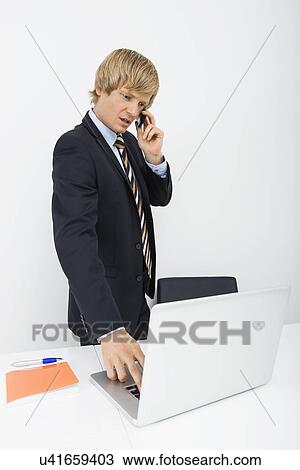 Mid adult businessman conversing on cell phone while using laptop in office View Large Photo Image Stock Image - Mid adult businessman conversing on cell phone while using laptop in office. Fotosearch