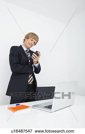 Mid adult businessman using cell phone at desk with laptop in office View Large Photo Image Stock Photo - Mid adult businessman using cell phone at desk with laptop in office. Fotosearch