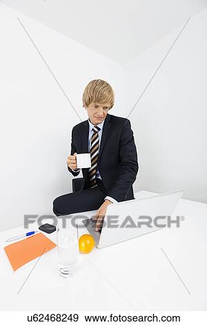 Mid adult businessman with coffee cup using laptop at desk in office View Large Photo Image Stock Photo - Mid adult businessman with coffee cup using laptop at desk in office. Fotosearch