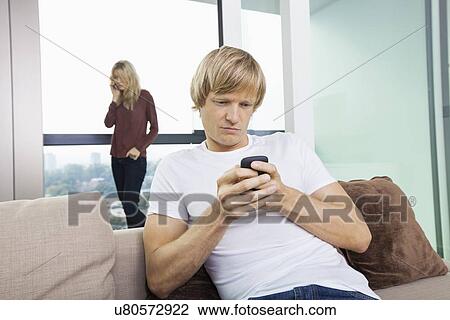 Mid-adult man text messaging while woman using cell phone in background at home View Large Photo Image Stock Image - Mid-adult man text messaging while woman using cell phone in background at home. Fotosearch