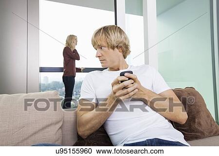 Mid-adult man text messaging while woman using cell phone in background at home View Large Photo Image Stock Image - Mid-adult man text messaging while woman using cell phone in background at home. Fotosearch