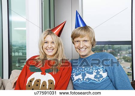 Stock Image - Portrait of cheerful couple wearing Christmas sweaters and party hats in living room at home. Fotosearch