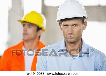 Stock Image - Portrait of confident male architect at construction site with coworker in background. Fotosearch