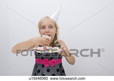 Stock Image - Portrait of cute girl eating birthday cake slice at table in house. Fotosearch