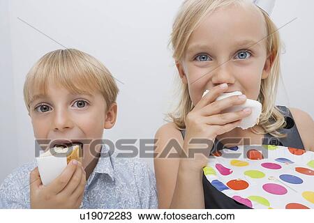 Stock Image - Portrait of cute siblings eating birthday cake slices in house. Fotosearch