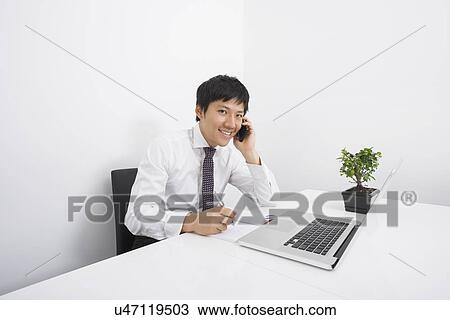 Stock Image - Portrait of happy businessman using cell phone while working at office desk. Fotosearch
