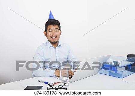 Portrait of happy businessman wearing party hat sitting at desk in office View Large Photo Image Stock Photo - Portrait of happy businessman wearing party hat sitting at desk in office. Fotosearch