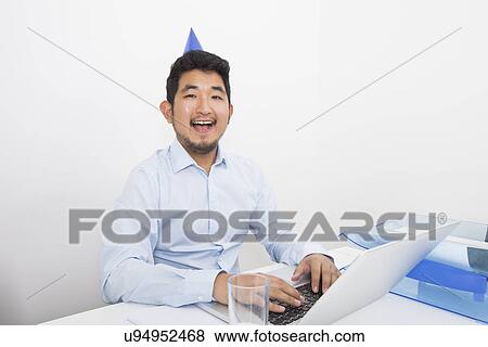 Stock Photo - Portrait of happy businessman wearing party hat while working in office. Fotosearch