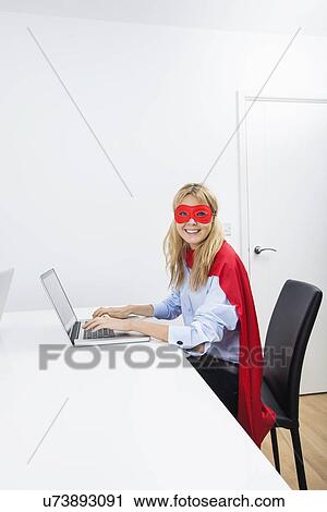 Stock Image - Portrait of happy businesswoman in superhero costume using laptop at office. Fotosearch