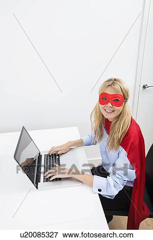 Stock Photo - Portrait of happy businesswoman in superhero costume working on laptop in office. Fotosearch
