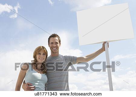 Stock Image - Portrait of happy couple with blank sign board against cloudy sky. Fotosearch