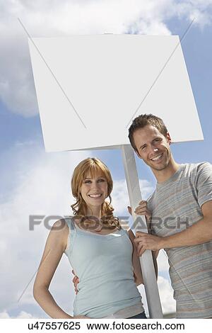 Stock Photography - Portrait of happy couple with blank sign over cloudy sky. Fotosearch