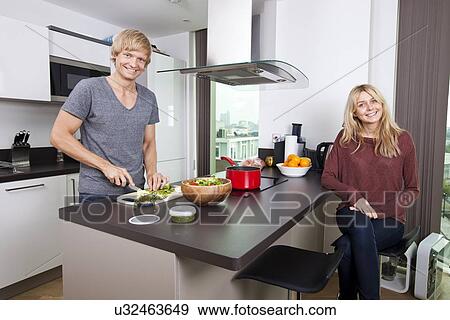 Stock Photo - Portrait of happy man cutting vegetables while woman sitting at kitchen counter. Fotosearch