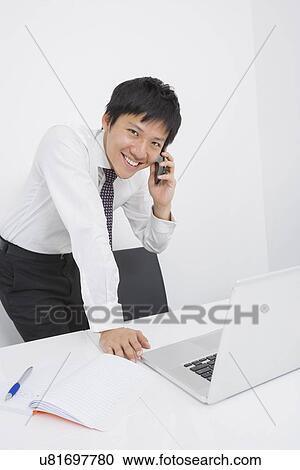 Stock Image - Portrait of happy mid adult businessman using cell phone at desk. Fotosearch