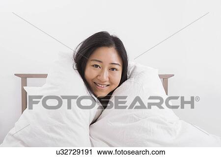 Stock Image - Portrait of happy woman wrapped in quilt on bed. Fotosearch