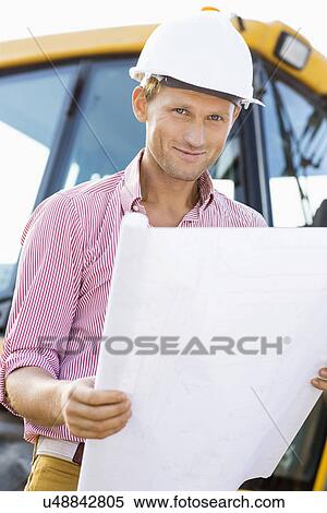 Stock Photography - Portrait of male architect holding blueprint at construction site. Fotosearch