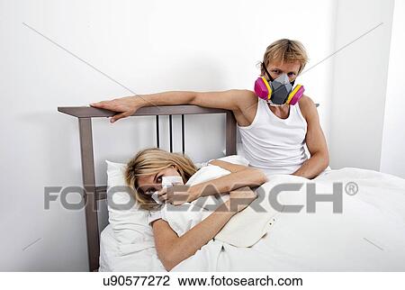 Stock Image - Portrait of man wearing gas mask while woman suffering from cold in bed. Fotosearch