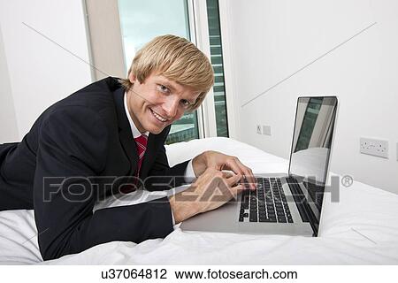 Stock Image - Portrait of mid adult businessman using laptop in bed at home. Fotosearch