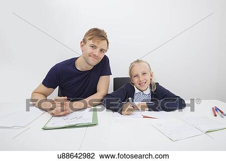 Stock Image - Portrait of mid adult man sitting with daughter studying at table. Fotosearch