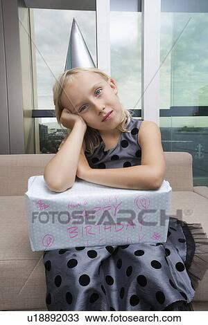 Stock Image - Portrait of sad girl with birthday gift sitting on sofa at home. Fotosearch