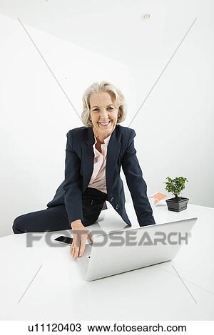 Portrait of senior businesswoman with laptop sitting on desk in office View Large Photo Image Stock Image - Portrait of senior businesswoman with laptop sitting on desk in office. Fotosearch