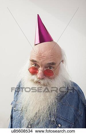 Picture - Portrait of senior man wearing red glasses and party hat against gray background. Fotosearch