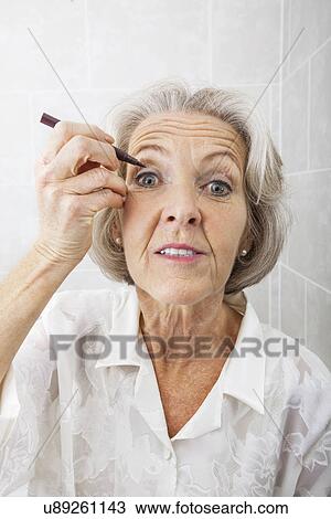 Stock Image - Portrait of senior woman applying eyeliner in bathroom. Fotosearch