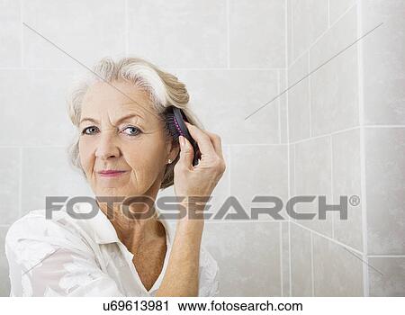 Stock Image - Portrait of senior woman brushing hair in bathroom. Fotosearch