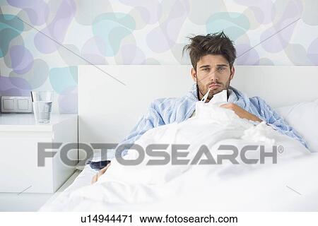 Stock Image - Portrait of sick man with thermometer in mouth reclining on bed at home. Fotosearch